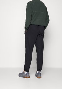 Muž stojící s rukama v kapsách, oblečený v černých teplákách, tmavozeleném strukturovaném svetru s dlouhým rukávem, bílých ponožkách a šedých teniskách s oranžovými podrážkami.