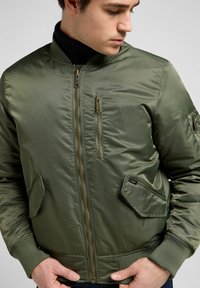 Giacca bomber verde oliva con finitura liscia, hardware dorato, tasche con zip, polsini a coste e colletto rialzato.