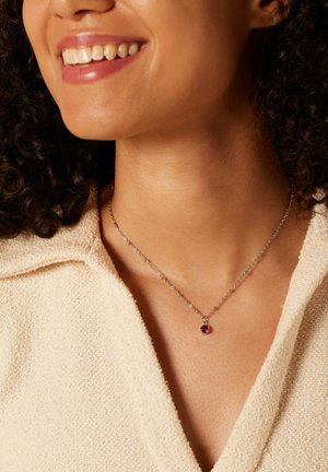 Glunderende vrouw die een fijne zilveren kettinkje draagt met een klein rond paarse hanger, gekleed in een crèmekleurige top met structuur.