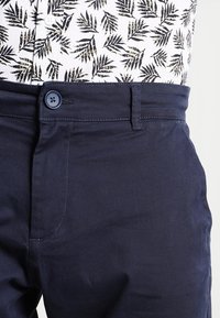 Laivastonsiniset housut, joissa on nappi ja vyölenkit, yhdistettynä valkoiseen paitaan, jossa musta lehtikuvio.