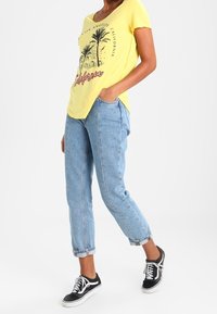 Gul t-shirt med V-ringning och palmträdsgrafik, i kombination med ljusblå, uppvikta jeans och svarta sneakers med vita snörningar.