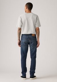 Gray short-sleeve t-shirt with a relaxed fit worn with dark blue denim jeans. Jeans feature a brown leather patch and classic back pocket design.