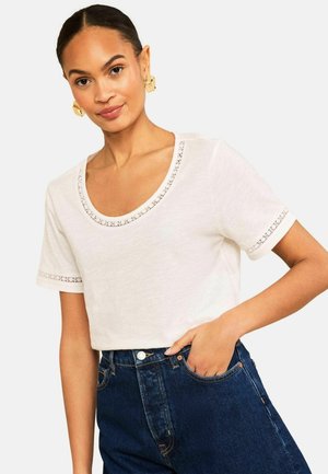 Nainen, jolla on kultaiset korvakorut, valkoinen lyhythihainen pusero pitsireunuksella ja tummansiniset korkeavyötäröiset farkut, käsi taskussa.