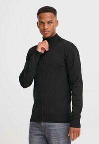 En ung man som bär en svart zip-up-tröja står självsäkert och greppar försiktigt kragen med sin högra hand, framför en neutral bakgrund.