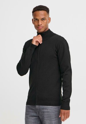 En ung mann iført en svart zip-up genser står selvsikkert og holder forsiktig tak i kragen med høyre hånd, foran en ensfarget bakgrunn.