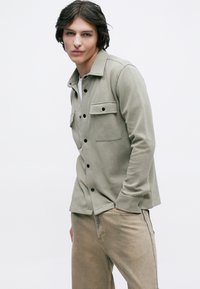 Νεαρός άνδρας με σκούρα μαλλιά, που φοράει ένα ανοιχτό γκρι σακάκι με κουμπιά και μπεζ παντελόνι, ποζάροντας μπροστά σε έναν απλό λευκό φόντο.