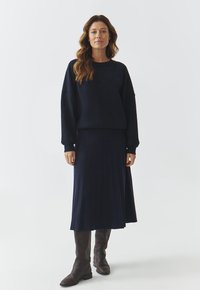 Sweatshirt marine avec épaules tombantes, associé à une jupe midi plissée assortie et des bottes brunes jusqu'aux genoux. Textures lisses tout au long.