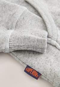 Grauer Pullover mit gerippten Bündchen und Kragen, aus strukturiertem Stoff, mit einem sichtbaren orangefarbenen und marineblauen Markenzeichen nahe dem Bündchen.