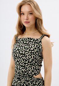 Schwarzes Blumenshirt mit kleinen weißen Gänseblümchen, quadratischem Halsausschnitt und geknoteten Trägern. Taillierte Passform mit Ausschnittdetail an der Taille.