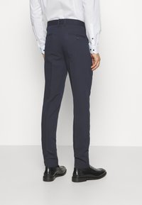 Marineblaue Hose mit geradem Schnitt, glatter Textur, Gürtelschlaufen und zwei Gesäßtaschen, kombiniert mit schwarzen formalen Schuhen.
