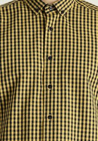 Camisa de cuadros amarilla y negra con cuello abotonado, presenta botones negros y una textura en la tela. Corte regular con mangas cortas.