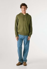 Sudadera verde oliva con un bolsillo frontal, puños y dobladillo de canalé; combinada con jeans azul claro y zapatos marrones con cordones.