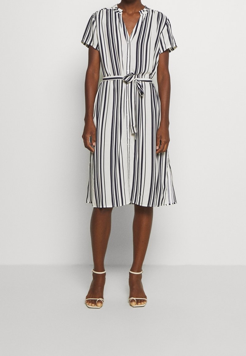 Femme portant une robe blanche mi-genoux avec des rayures noires verticales, des manches courtes, une fermeture à boutons à l’avant et une ceinture nouée à la taille.