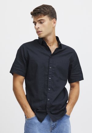 Koszula krótko rękawowa z button-up w ciemnoniebieskim materiale, z kołnierzykiem, kieszenią na piersi oraz czarnymi guzikami. Noszona z rękami w kieszeniach.