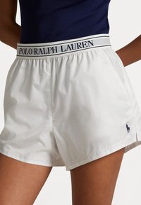 Vita shorts gjorda av mjukt, lätt tyg. Har en marinblå logotyp och ett elastiskt midjeband med "POLO RALPH LAUREN" i marinblå text.
