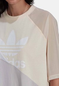 T-shirt oversize crema e beige chiaro con logo Adidas bianco. Presenta un mix di materiali opachi e trasparenti, maniche raglan e una vestibilità rilassata.