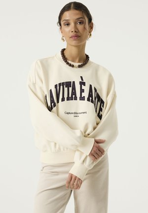 Vrouw met een crèmekleurige sweatshirt met de tekst “LA VITA È ARTE”, beige broek, een grove bruine ketting en gouden oorringen, staat rustig.