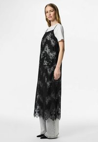 Sieviete tērpusies melnā mežģīņu naktskrekla stilā pār baltu T-kreklu un gaiši pelēnām džinsām, stāv sāniski ar rokām atpūstā pozīcijā gar sāniem.