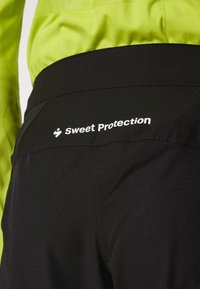 Sorte bukser med en glat tekstur, med en synlig hvid logo "Sweet Protection" på bagsiden af taljebåndet. Letvægtsstofdesign.