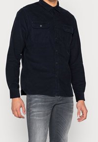 Laivastonsininen puuvillakonnapaita, jossa on nepparikiinnitys, kaksi etutaskua, pitkät hihat ja klassinen kaulus. Sileä pinta ja hienovarainen kiilto.