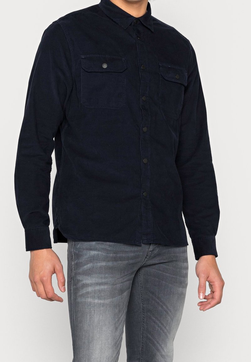 Marineblauw corduroy overhemd met knopen, voorzien van twee borstzakken, lange mouwen en een klassieke kraag. Gladde textuur en subtiele glans.