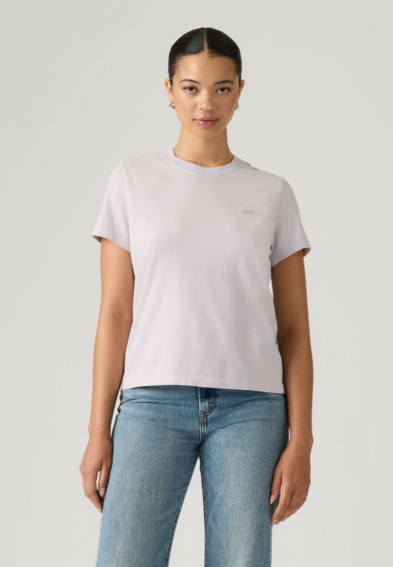 Femme aux cheveux foncés portant une chemise à manches courtes violet clair et un jean bleu, debout devant un fond clair uni.