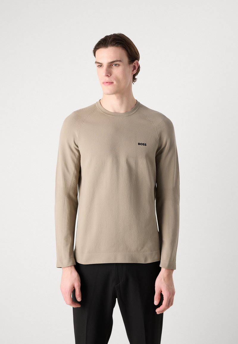 Camisa de manga larga beige con cuello redondo, tejido texturizado y logo negro de "BOSS" en el lado izquierdo del pecho, combinada con pantalones negros.
