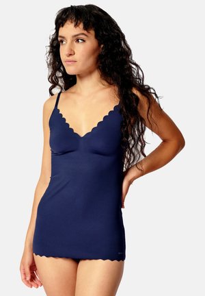 Woman with long curly hair wearing a fitted navy blue camisole with scalloped edges, standing with one hand on her hip.