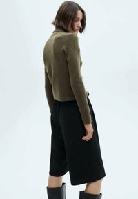 Olijfgroene cropped coltrui van zacht tricotmateriaal, gecombineerd met zwarte knielange shorts met een relaxte pasvorm en een tailored ontwerp.