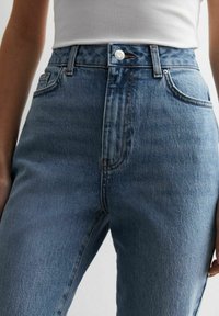 Vaqueros de mezclilla azul de tiro alto con una textura suave, ligero desvanecimiento, diseño clásico de cinco bolsillos y cierre con botón plateado.