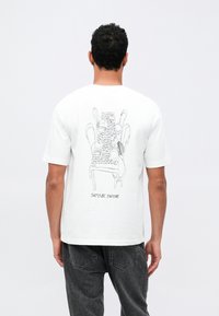 Hombre vistiendo una camiseta blanca con un arte lineal negro de sillas apiladas y texto manuscrito en la parte posterior, combinada con jeans oscuros.