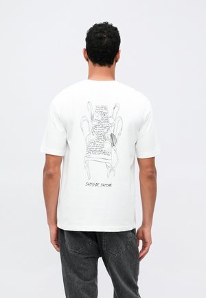 Vīrietis, kurš valkā baltu T-kreklu ar melnu līniju mākslu, kas attēlo sakrautas krēslus, un rokrakstā rakstītu tekstu uz muguras, apvienots ar tumšām džinsām.