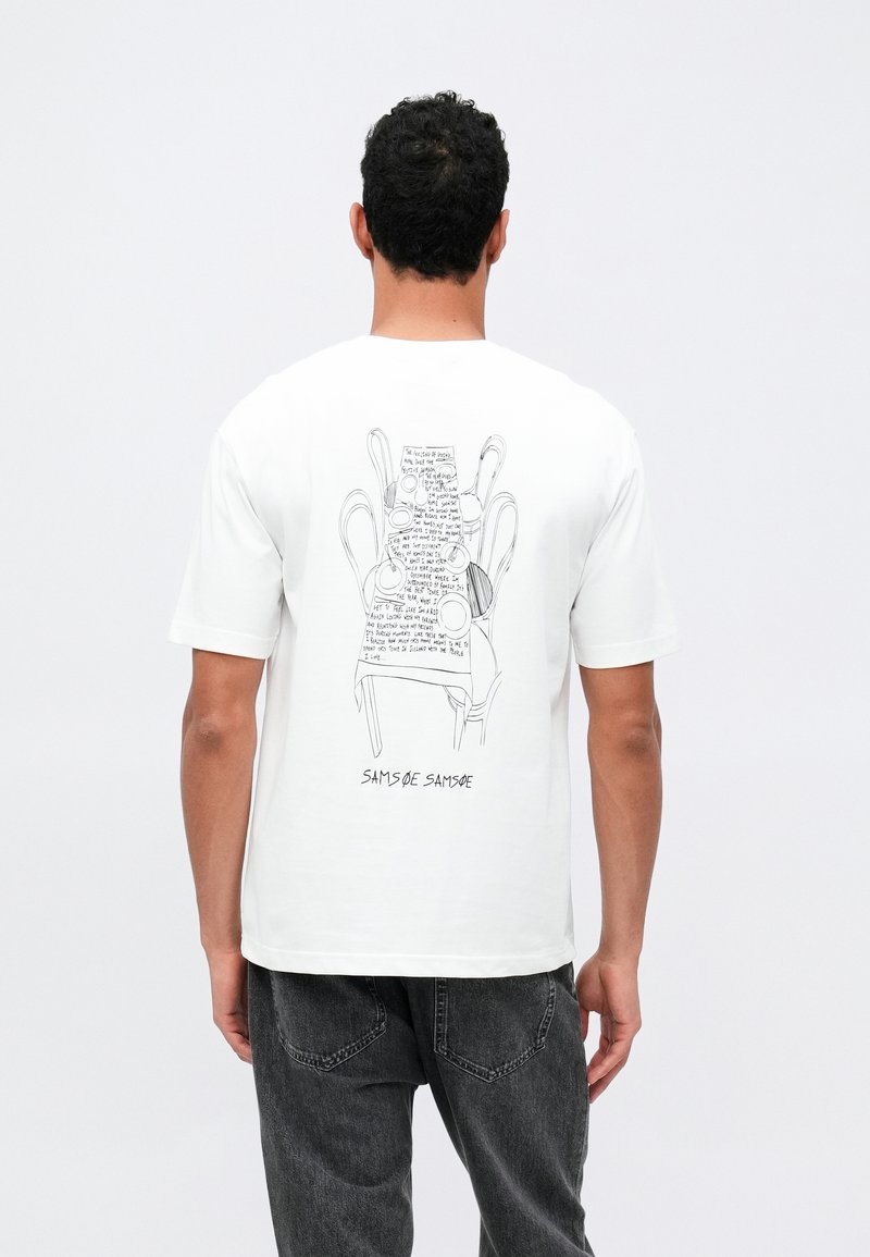 Vīrietis, kurš valkā baltu T-kreklu ar melnu līniju mākslu, kas attēlo sakrautas krēslus, un rokrakstā rakstītu tekstu uz muguras, apvienots ar tumšām džinsām.