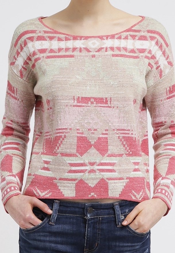 Žena v růžovém a béžovém vzorovaném svetru s dlouhým rukávem, který má geometrické tvary, a modrých džínách, s rukama v kapsách.