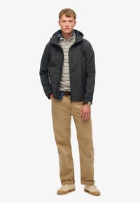 Męska czarna kurtka softshell z kapturem, szary polo w paski, beżowe chinosy i kremowe sneakersy. Posiada zamek błyskawiczny z przodu i boczne kieszenie.