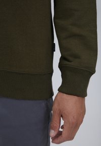 Oliven grønn sweatshirt med ribbestrikkede mansjetter og linning. Struktureret stoff, tettsittende design, med logoetikett på siden.