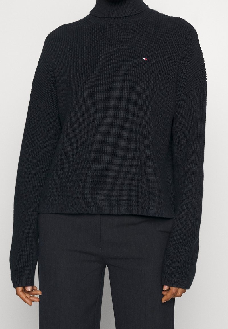 Persona che indossa un maglione nero a collo alto a coste con maniche lunghe e pantaloni scuri su uno sfondo semplice e chiaro.