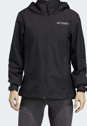 Sort hættejakke med fuld lynlås og TERREX-logo på brystet, båret sammen med mørke leggings med mønster, hænder afslappet ved siden.