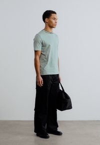 Ανοιχτό πράσινο κοντομάνικο μπλουζάκι με μαύρο γιακά, συνδυασμένο με μαύρο cargo παντελόνι φαρδιάς γραμμής και υφασματινή μαύρη τσάντα με υφή. Ουδέτερο φόντο.