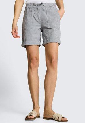 Kobieta ubrana w szare szorty w paski z podwiniętymi brzegami i beżowe klapki na nogach, stoi z jedną ręką w kieszeni.