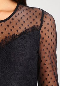 Black top featuring a sheer dotted mesh upper and a textured lace lower section, with a round neckline and long sleeves.