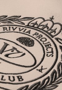 Bež tkanina s crnim vezenim okruglim logotipom s ukrasnim elementima, tekstom i teksturiranim dizajnom krune i listova.