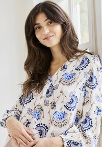 Jeune femme aux longs cheveux bruns portant un chemisier blanc avec des motifs floraux bleus et beiges, debout près d'une fenêtre avec une lumière naturelle douce.