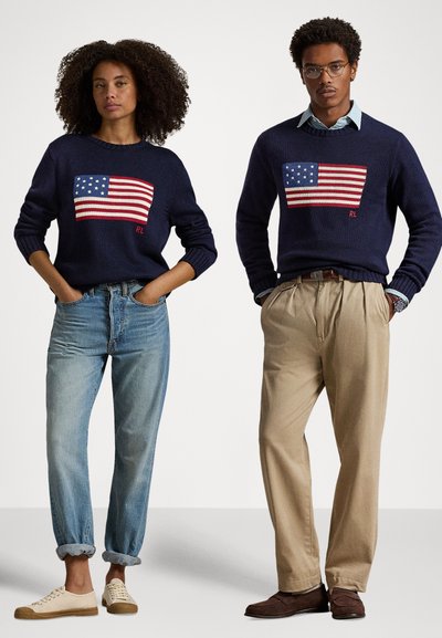 Námořnické svetry s pleteným motivem americké vlajky. Žena má na sobě modré džíny a krémové tenisky; muž má na sobě khaki kalhoty a hnědé mokasíny.