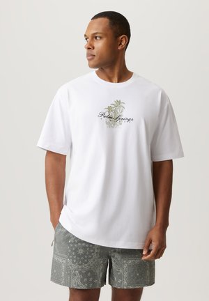 Biała bawełniana koszulka z nadrukiem palmy i tekstem "Palm Springs" w kolorze czarnym, w połączeniu ze wzorzystymi szarymi szortami.