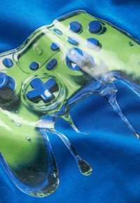 T-shirt graphique avec un design de manette de jeu verte sur fond bleu, avec des détails brillants et des gouttes texturées ressemblant à du liquide.
