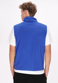 Blaues Fleece-Weste mit hohem Kragen, glatter Textur und ohne Verschlüsse, getragen über einem weißen Kurzarmhemd.