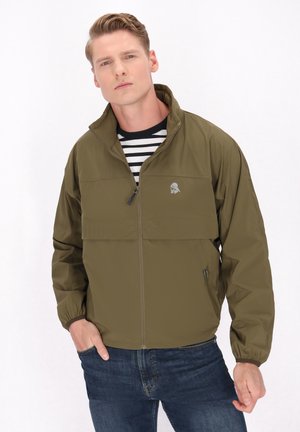 Jachetă de culoare verde măslin cu design cu jumătate de fermoar, manșete elastice și un mic logo pe piept, asortată cu o cămașă cu dungi și blugi albaștri.