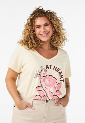 Smilende kvinde med krøllet hår iført cremefarvet skjorte med en lyserød blomst og teksten "Wild at Heart", hænderne i lommerne på lyse beige bukser.