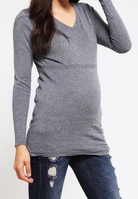 Graues V-Ausschnitt Langarmshirt aus weichem Material, figurbetonte Passform mit einem dehnbaren Bund, kombiniert mit abgetragenen blauen Jeans. Keine auffälligen Muster oder Akzente.